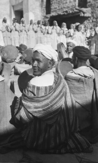 A Berber musician seated on the floor with a tambourine in his hand and looking back at the photographer over his left shoulder. He is wearing a white turban and a striped Djellaba. A dance troupe can be seen in the background. Telouet, 13th January 1936.