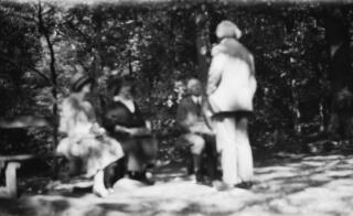 David & Margaret Lloyd George with another couple in a woodland clearing. D Ll G is standing, the others sitting, as though listening to him. Out of focus.