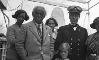 David & Countess Lloyd George in the middle distance walking arn in arm, their backs to the camera,along a track lined with tall trees on either side.
