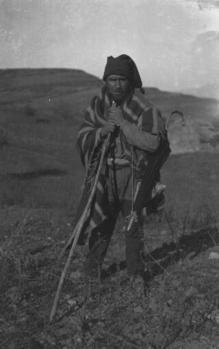 A shepherd carrying a long stick with an ancient umbrella slung over his left shoulder.