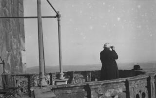Lloyd George, his back to the camera and standing on a balcony, looking through binoculars at the vista below.