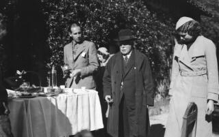 David Lloyd George in hat and coat, cigar in right hand, in the company of a young lady, a well-dressed younger man looking on.