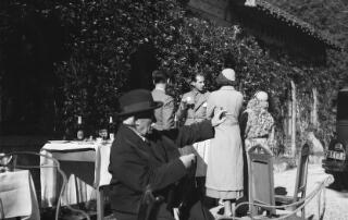 David Lloyd George sitting outside and wearing a hat and coat. He is gesturing with his left hand, as though talking to someone outside the photo. A bottle of 'Perola' beer is on a nearby table. This is a Portuguese beer, suggesting this is of his visit to Portugal in 1934.