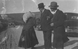 Two smartly dressed men addressing a peasant woman with a sack over hershoulder and her back to the camera. Behind them are cars presumed to be of David Lloyd George's entourage