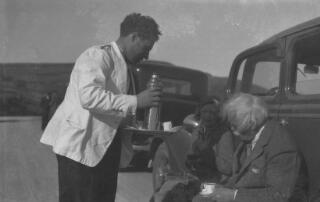 David & Megan Lloyd George sitting on the running board of a car, a waiter serving them with drinks from a thermos flask.