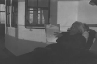 Head and shoulders informal portrait of David Lloyd George wearing a coat and sitting on a recliner on the deck of a ship reading papers, a cigar in his mouth.