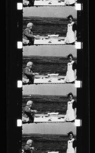 Four frames from a cinefilm showing David Lloyd George seated at an alfresco restaurant table. A smiling waitress is in attendance. Beyond them is a plain dotted with trees and cultivated fields.