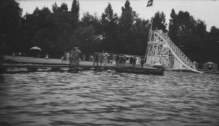 A small paved area adjacent to the lake. Visible is a waterslide and a small boat with 'Lausanne Clichy Plage' painted on the side. Image blurred probably due to camera shake.