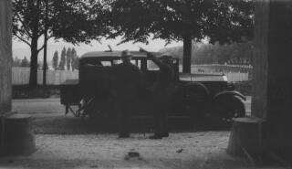 David Lloyd George standing beside his car in conversation with a man in a flat cap. Both are pointing in the direction in which the car has just come from.