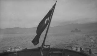 View from the stern of a boat showing the Red Ensign, a distant coastline and a twin funnelled steamer in the middle distance.
