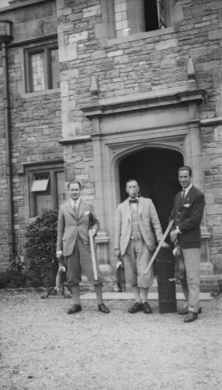 A J Sylvester wearing plus fours and carrying a shotgun and a dead rabbit standing in front of a doorway with two other men, one also carrying a shotgun. Both are holding dead rabbits. PZ7890/41 is a variant of this.