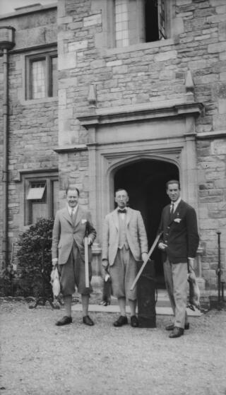 A J Sylvester wearing plus fours and carrying a shotgun and a dead rabbit standing in front of a doorway with two other men, one also carrying a shotgun. Both are holding dead rabbits. PZ7890/41 is a variant of this.