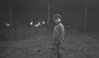 A young child wearing a beret and raincoat in a farmyard. Chickens are visible behind the child.