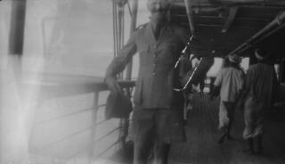 Torso and lower part of the face of a man in military uniform holding a topi and smiling. He is leaning against the hand rail of a ship. The presence of two men in traditional Indian costume suggests this may date from Lloyd George's visit to Ceylon (Sri Lanka) in 1931.