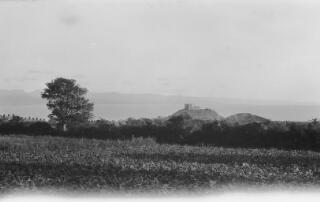 Cricieth Castle in the middle distance photographed from a field. The Merionethshire coastline is visible in the distance.