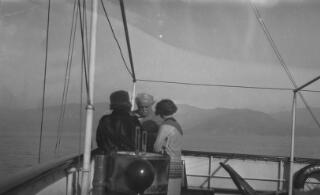 David & Margaret Lloyd George on the deck of 'Sabrina' in conversation with Megan. The coastline of France or Italy is visible in the distance.