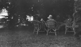 David, Margaret, Gwilym Lloyd George & others seated on cane chairs in the shade of a tree in a garden.