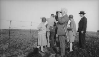 Members of the Lloyd George entourage standing behind a barbed wire fence. Gwilym is pointing at something in the distance, David Lloyd George, nearest the camera, is looking through binoculars. Very similar to PZ7890/47.