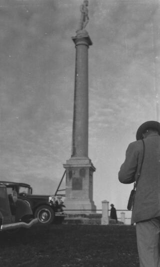 A statue of an unidentified figure on top of a tall column. An unidentified individual can be seen photographing the monument in the bottom right hand corner.