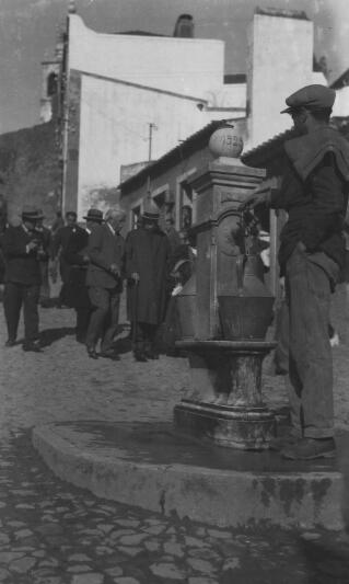 A man draws water from the village tap as Lloyd George and members of his enturage pass by.